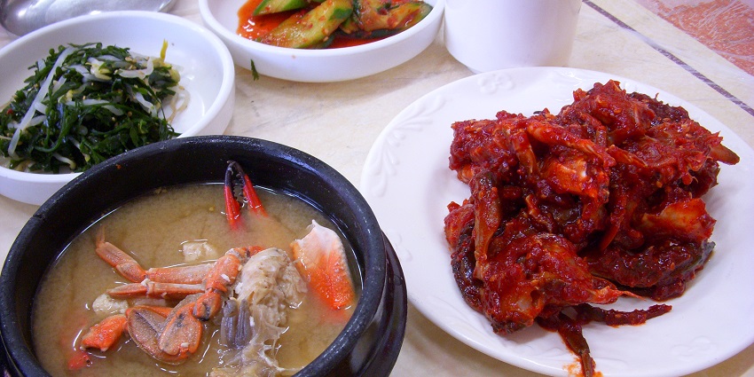 A table with a Korean meal, featuring a black bowl of crab stew, side dishes of vegetables