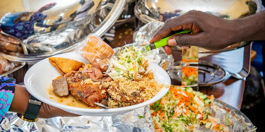 A person serving food from chafing dishes onto a plate containing regular Nigerian cuisine