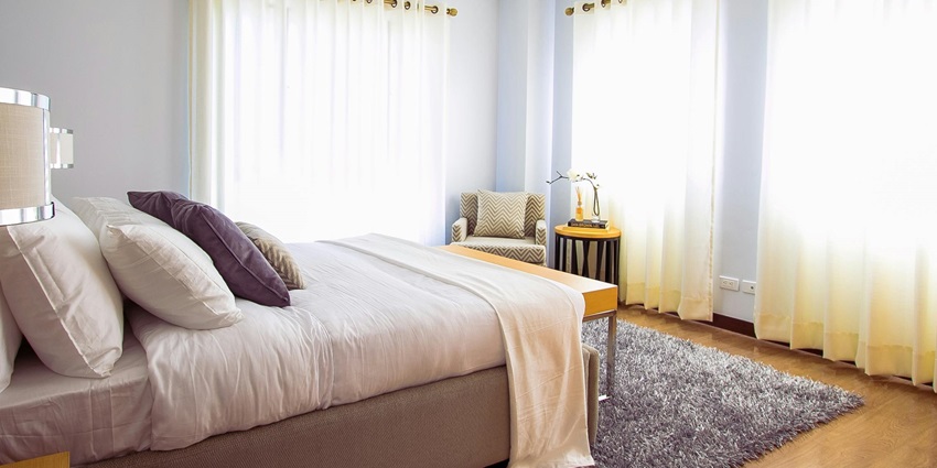 Room with a made bed and pillows on it and a chair in the corner and white curtains