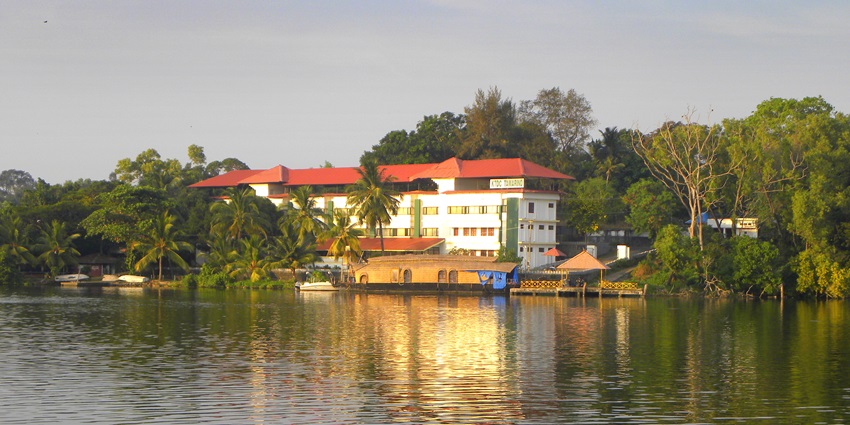 Tamarind KTDC easy hotel with a red roof and a houseboat parked surrounded by greenery.

