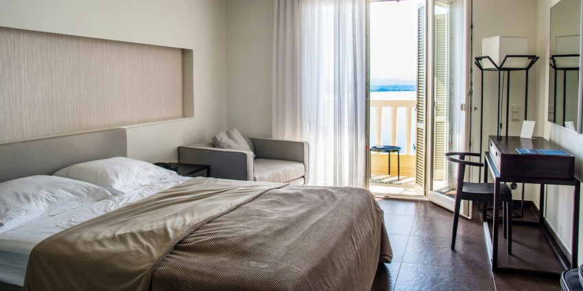 A hotel room with a neatly made bed, a balcony overlooking the ocean, and a desk