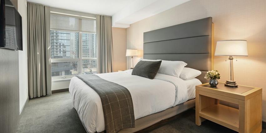 A hotel room with a neatly made bed, a headboard, and a window overlooking a cityscape