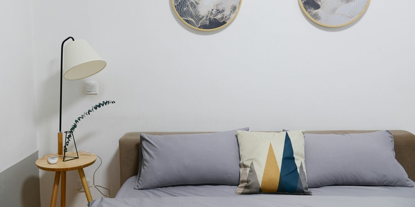 An image showing a beige wooden nightstand with a white desk lamp beside a cosy hotel bed.