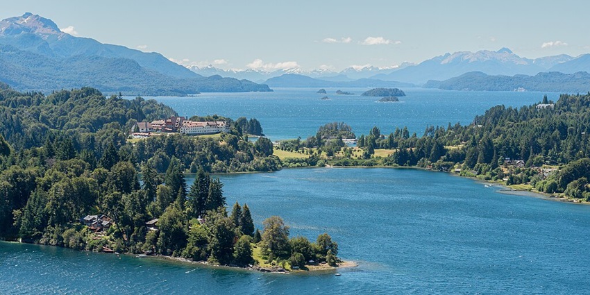 An aerial view of a huge lake with a few houses on the left surrounded by greenery and hills.