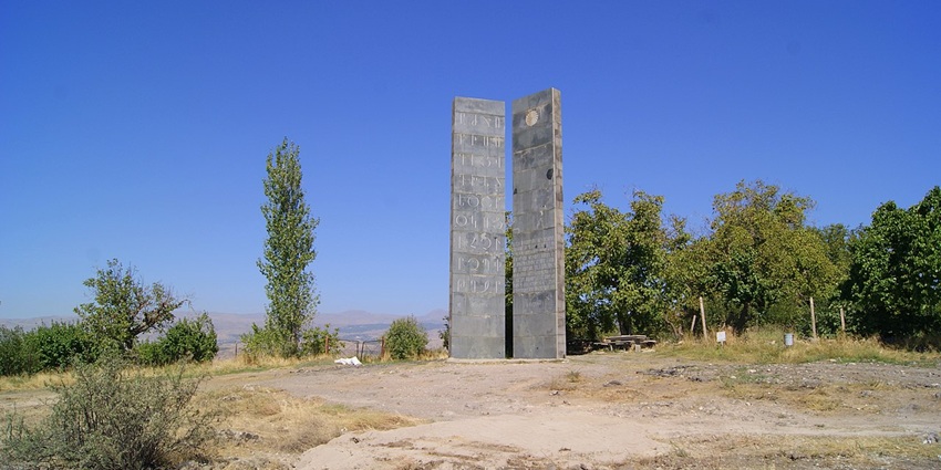 A frontal view of two tall stone pillars with the Armenian alphabet written all over it, surrounded by greenery