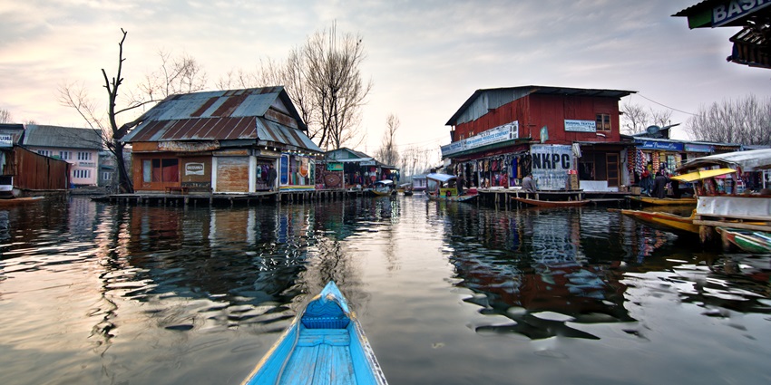 Floating market on the water with multiple shops, a blue boat and dry trees in the back