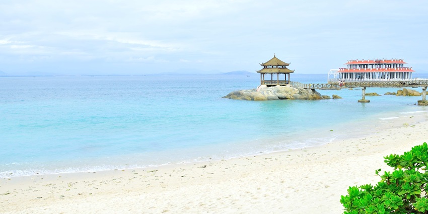 Wuzhizhou Island, a beach with clear turquoise water, a gazebo on a rock, and a building