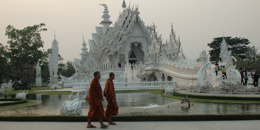 Intricate architecture of the Wat Rong Khun temple with two monks walking in the front of it