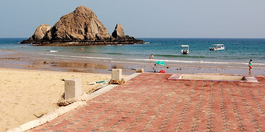 Rocky island offshore, people swimming and walking along a sandy beach with boats nearby.