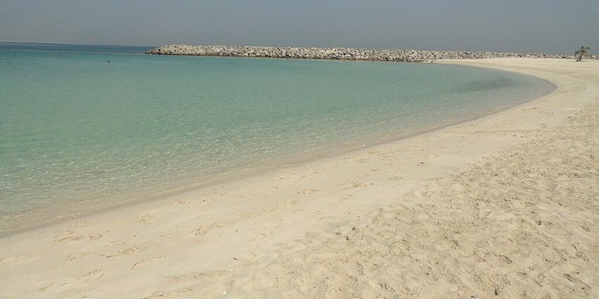 A serene beach with clear turquoise water, a stone breakwater, and a calm, cloudless sky.