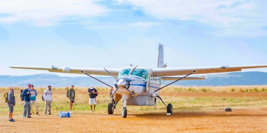 Passengers standing next to an aeroplane on sandy ground, distant green plains and hills.