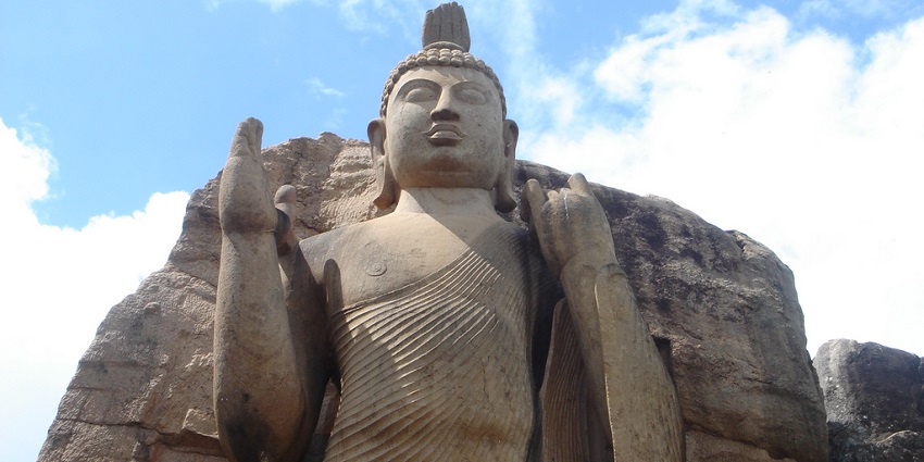 Ancient Aukana Buddha statue carved from stone, standing tall against a blue sky.