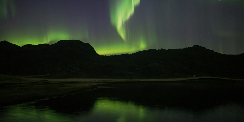 An image showing a dark landscape illuminated by a bright green Aurora Borealis, like in Aurora Reykjavík