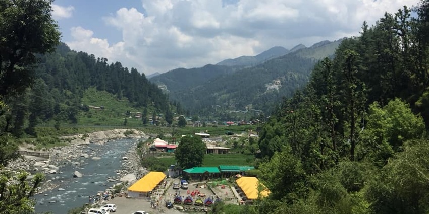 An image of yellow and green tents by a river, in the Barot valley with green hills.