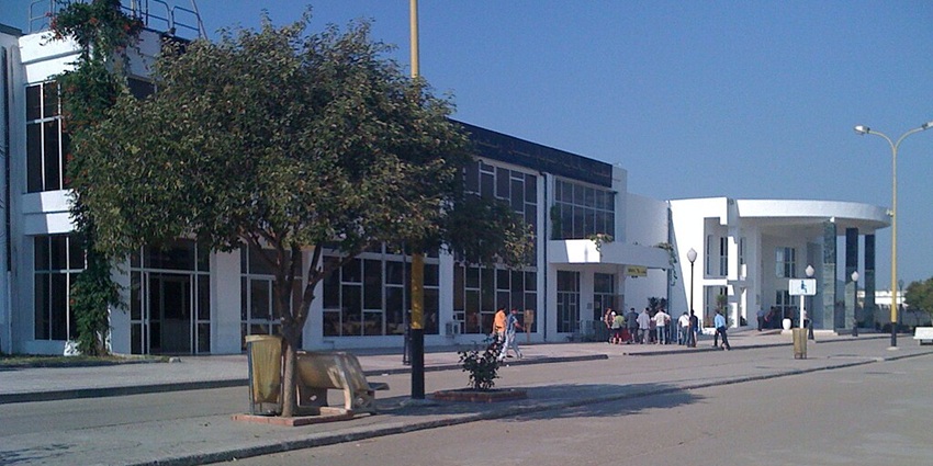 People walking outside a modern building with large glass windows, trees, and clear skies.