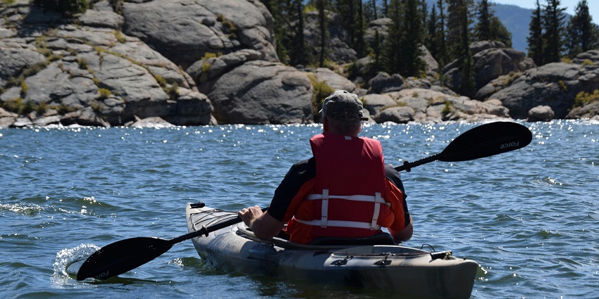 Person wearing safety vest kayaking and holding a paddle with rocky shoreline and trees