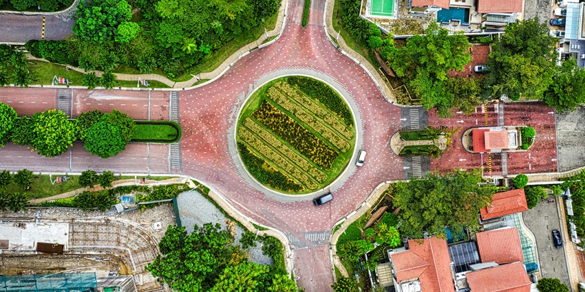 An image of a landscaped roundabout with greenery, red-roofed buildings, a nearby tennis court, and urban roads