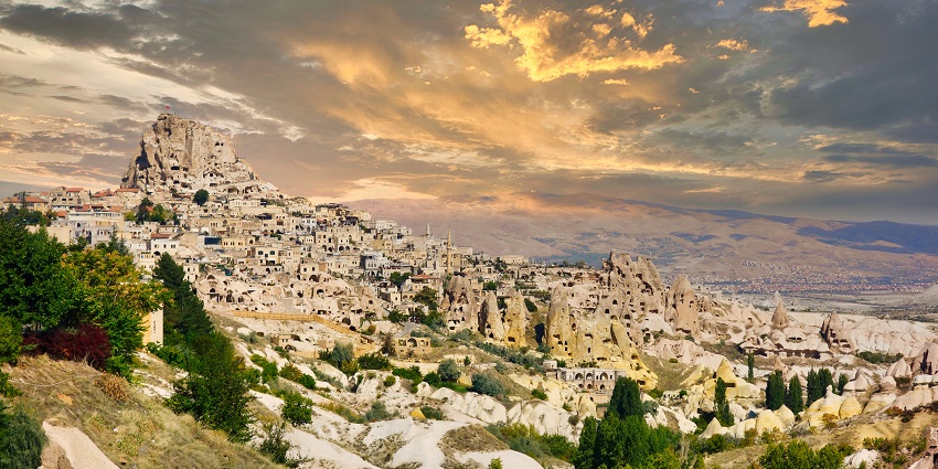 An image of the Uchisar Castle illuminated by golden hues during sunset, with the Cappadocian valleys in the background.