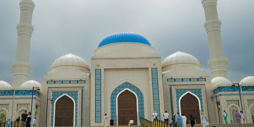 People walk toward a grand mosque with blue domes and tall minarets under a cloudy sky.