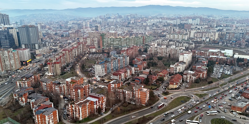 Urban landscape with buildings, busy roads, and distant mountains under a hazy sky.