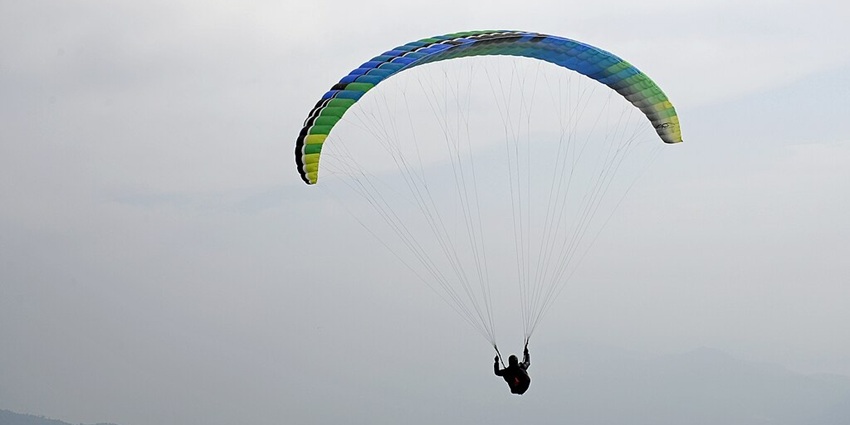 An image of a person paragliding in Bir Billing, under a large, colourful parachute above hazy mountains and valleys.