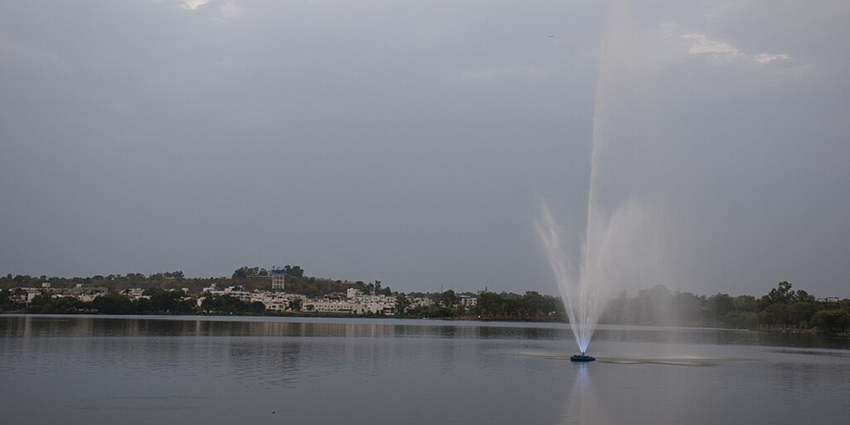 Lake with a fountain in the middle of the water body and distant buildings and greenery