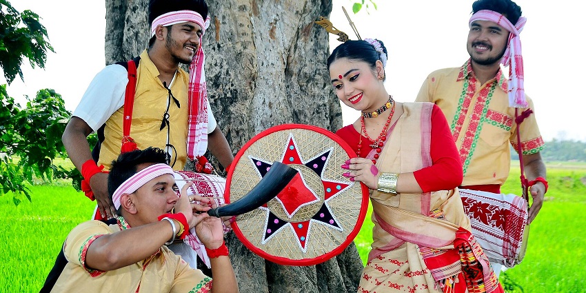 An image of Assamese people in traditional attire celebrating Bihu with music, dance, a buffalo horn, and a jaapi.