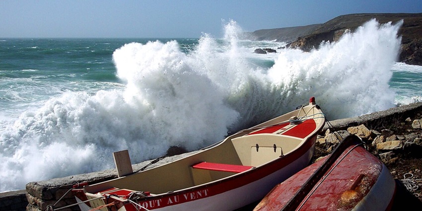 Powerful ocean waves crash against rocky cliffs as small boats rest on the rugged shore