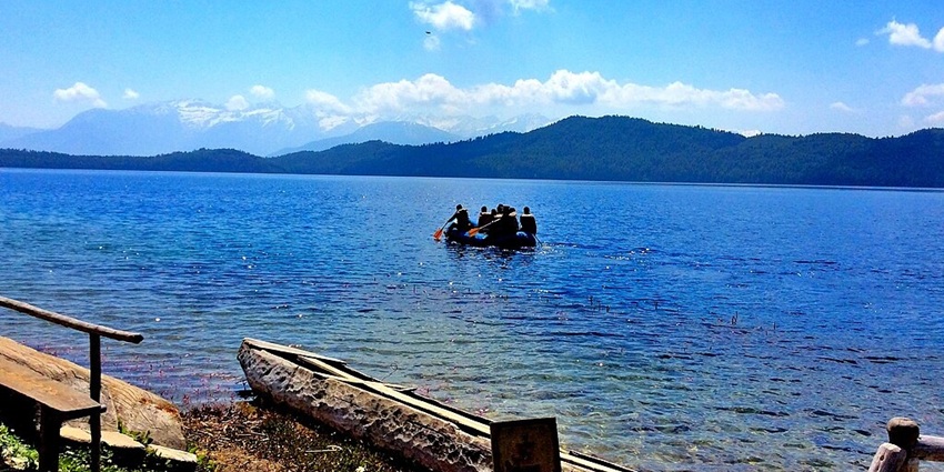 People boating with paddles against the waters of Rara Lake in Nepal with distant mountains.