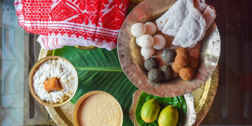 An image of traditional attire and a food thali celebrating Bohag Bihu, with one playing a buffalo horn instrument, the other dancing
