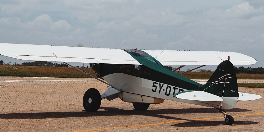 A white small propeller aircraft parked on an airstrip with big wheels and greenery