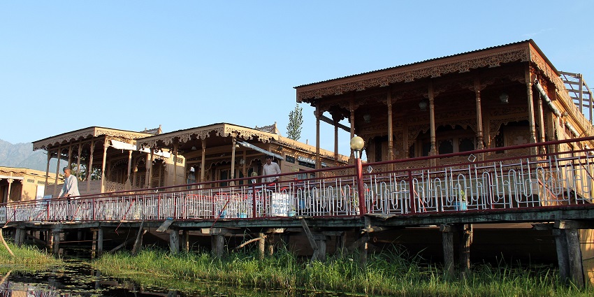 Multiple houseboats with people standing near the houseboats and water weeds in the lake.