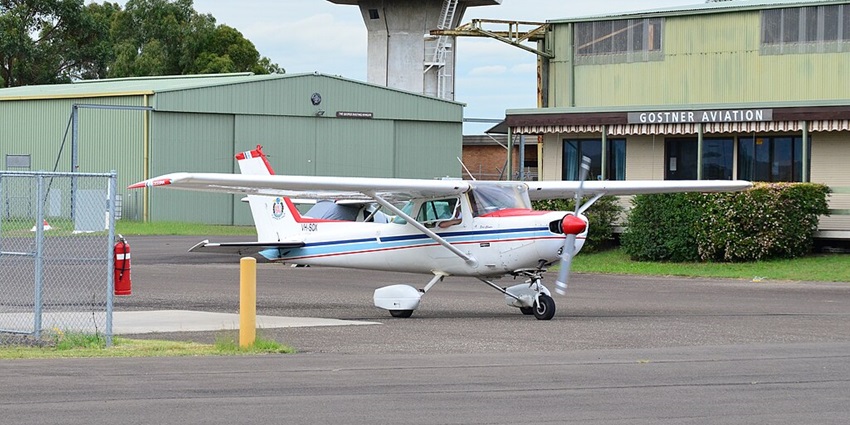 A small aircraft is taxiing at the Camden Airport beside hangars and control tower.