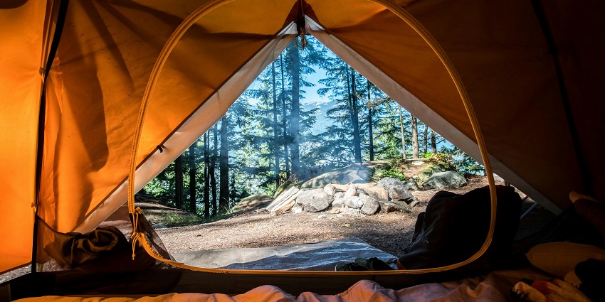 View from inside a tent looking out at a campfire, trees, and mountains on a sunny day.