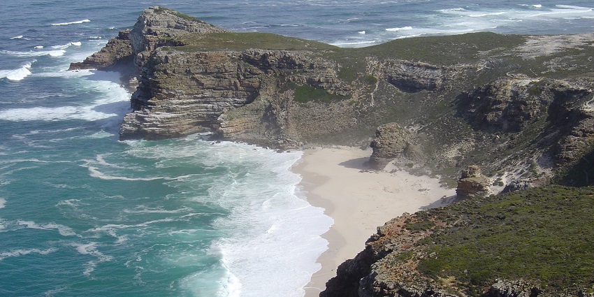 Rugged cliffs and sandy beach by ocean waves at the Cape of Good Hope, South Africa.