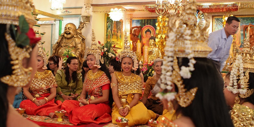 An image of a traditional ceremony with participants wearing ornate costumes, gold headpieces, floral arrangements, chandeliers.