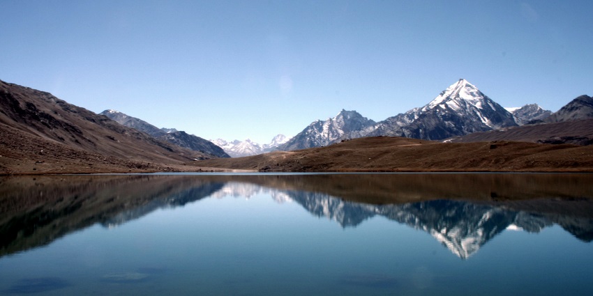 Chandratal Lake’s crescent-shaped shoreline encircles calm, reflective waters amid views.