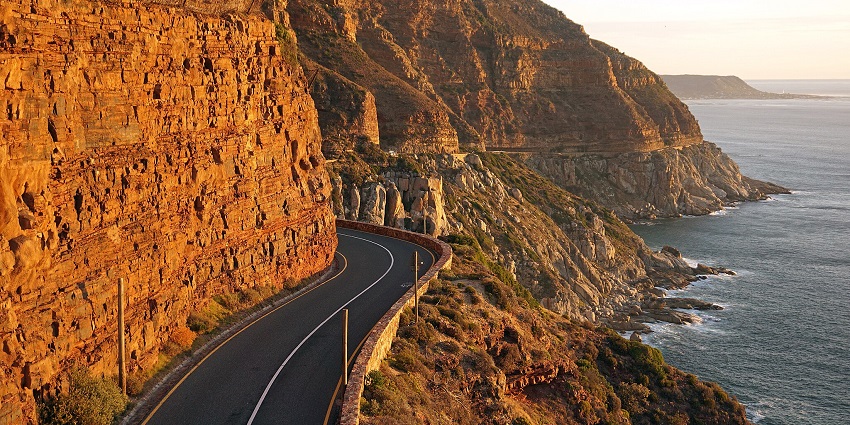 A view of the steep cliffside of Chapman’s Peak Dive, with a winding road bordered by stones.