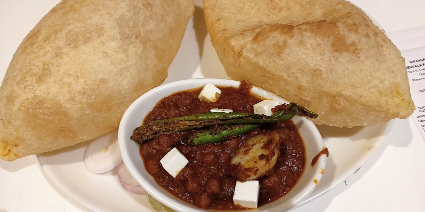 An image of Chole Bhature with two puffed breads, spicy chickpea curry, red onion slices, and pickle