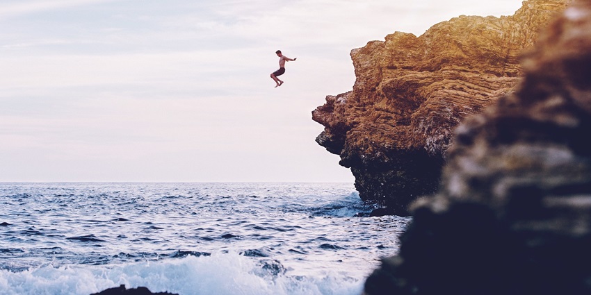 A man diving off a rocky cliff into the sea, with waves crashing under a cloudy sky.