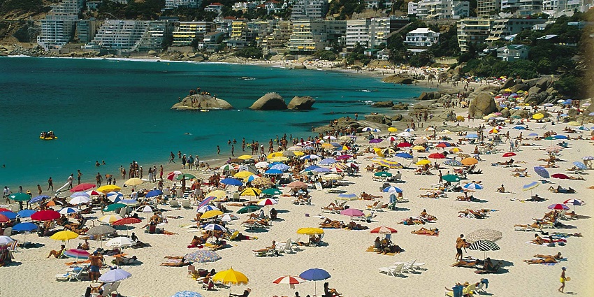 An image of a crowded Clifton beach, featuring colourful umbrellas, sunbathers, swimmers, clear blue waters, and rocky coastline.