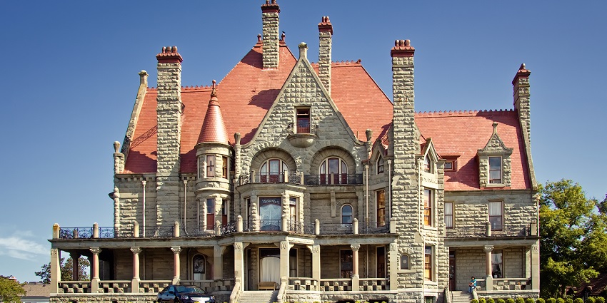 An image of a grand Victorian mansion of Craigdarroch Castle, with red, peaked turrets, intricate details, lawn, and shrubs.