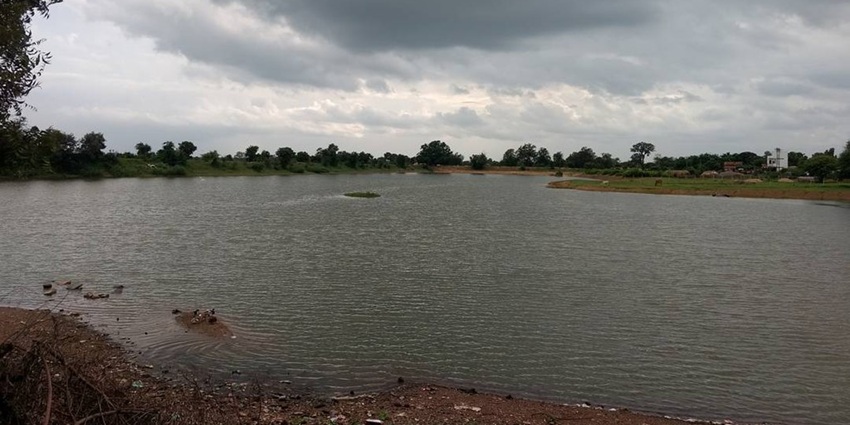 Lake with wet dirt shorelines surrounded by greenery and trees under a grey cloudy sky