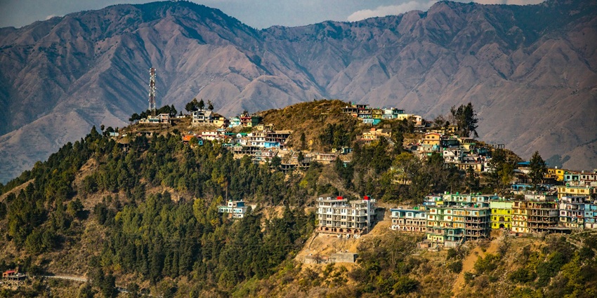 A serene view of Dhanaulti featuring towering peaks, a colourful town, and greenery.