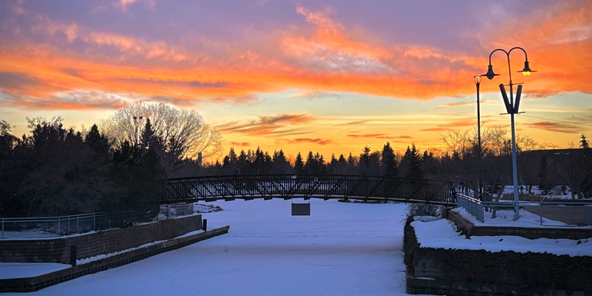 A snow-covered park with trees and streetlights. Sunset takes place at the backdrop.