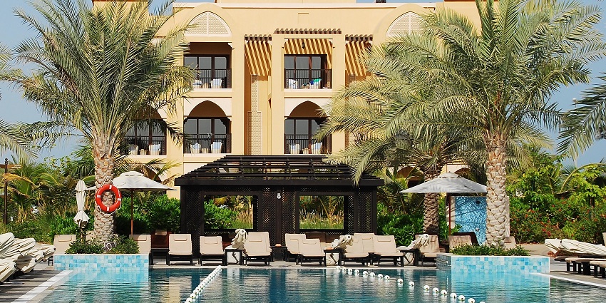 A hotel building with arched windows overlooks a swimming pool lined with lounge chairs