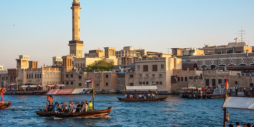 Boats with passengers sail along Dubai Creek, with traditional buildings and a minaret