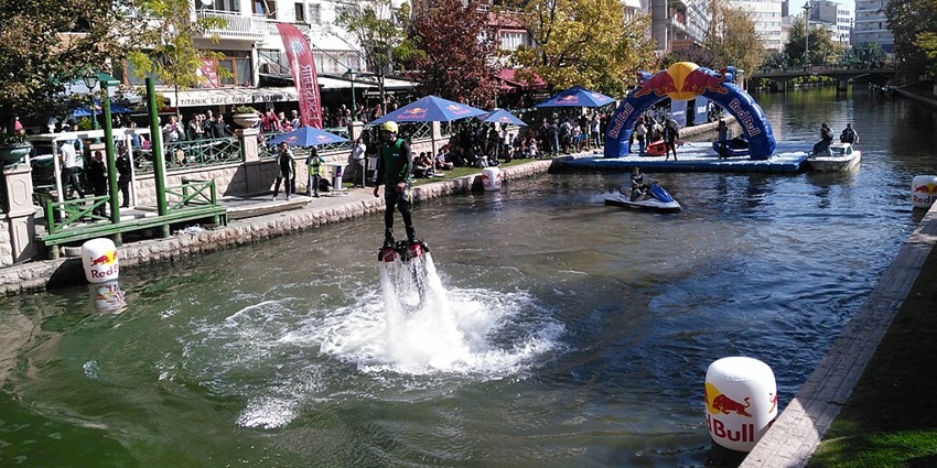A person flyboarding on a canal during a event, with crowds watching from both sides.