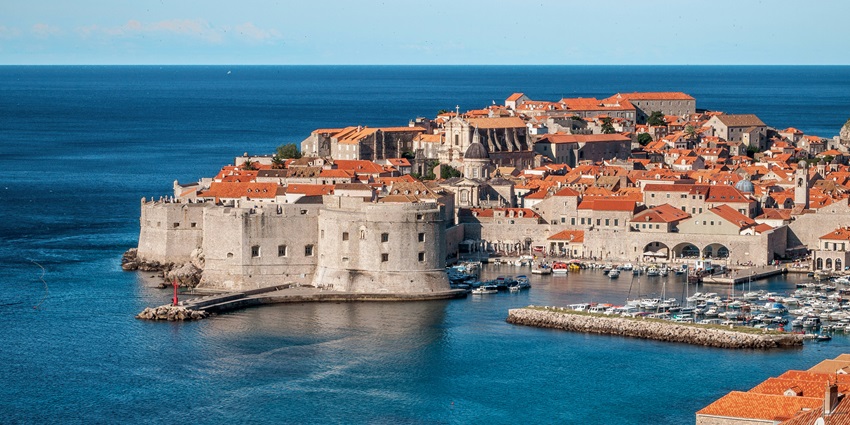 Tourists stroll along a scenic walkway between an ancient fortress and the stunning sea.