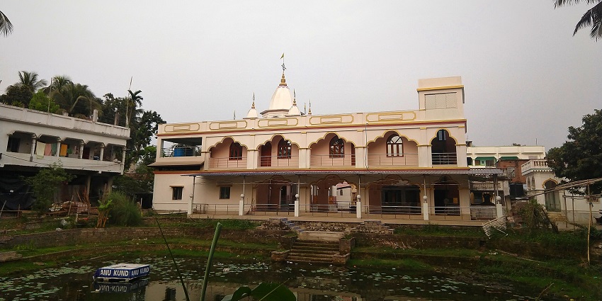 A temple with traditional architecture, a flag on top of the temple & a small water body.